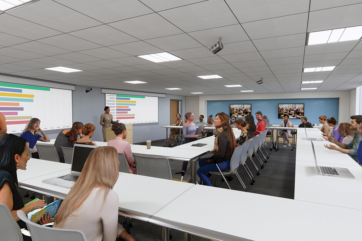 A classroom full of students are viewing projected slides of graphs while a woman stands at a podium at front of the classroom.