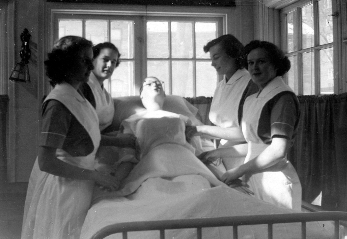 An old black and white photo from the early twentieth century shows 4 nursing students in white nursing uniforms stand around an artificial patient in a bed.