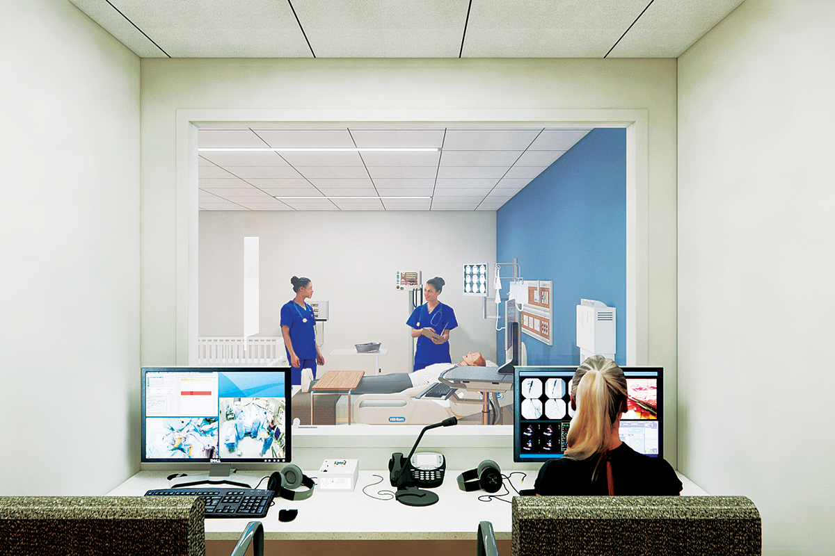 Two nursing students in blue scrubs attend to an artificial patient in a bed in a simulated isolation ward. Another looks on from a control room with several computer monitors in the foreground.