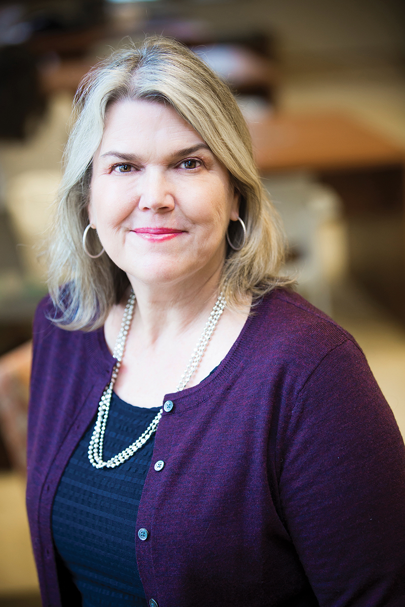 Lisa Thompson wears a purple sweater over a navy blue top in an environmental portrait.